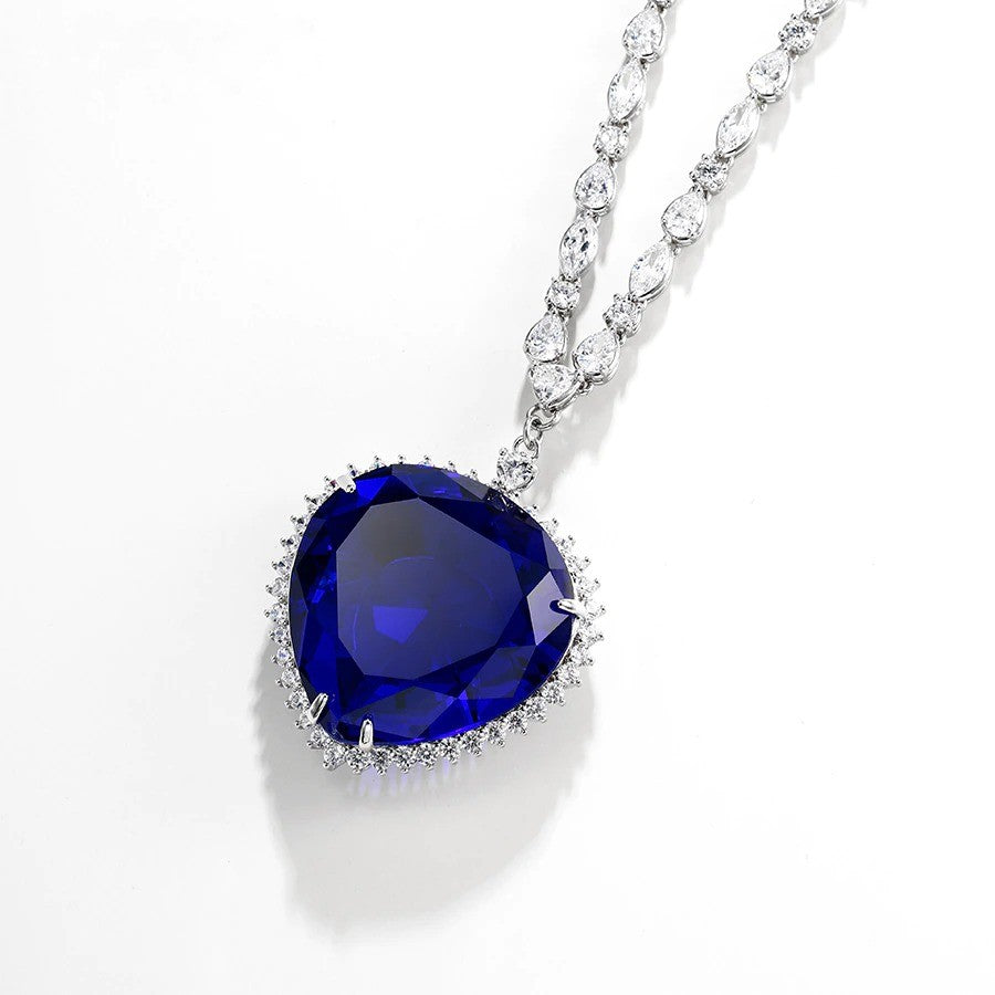 The heart of the ocean necklace replica from the Titanic movie. The pendant is a deep blue surrounded by small gems and is attached to a chain with clear, sparking gems. The necklace sits on a white background.