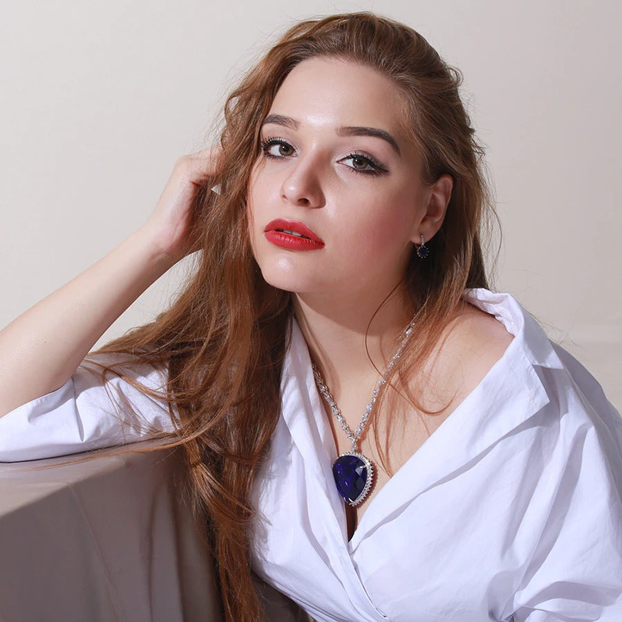 A woman is wearing a replica of the Heart of the Ocean necklace which features in the 1997 movie, Titanic. She is wearing a crisp, white collared shirt and has one hand in her hair.