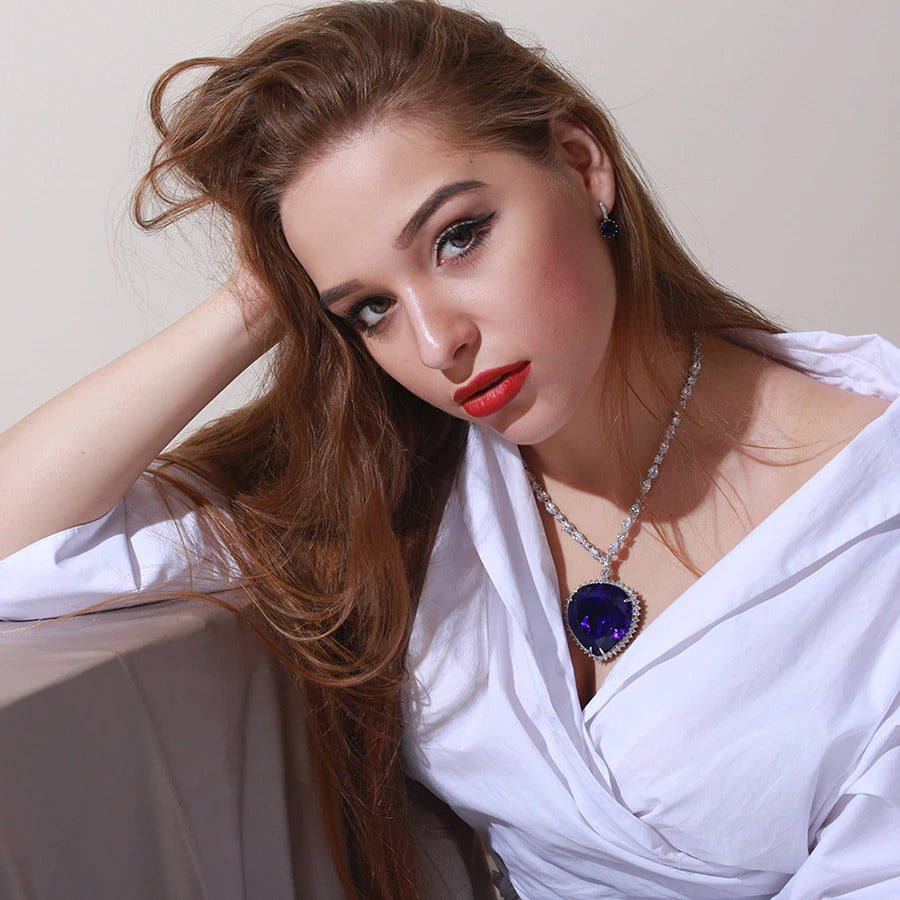 A woman is wearing a replica of the Heart of the Ocean necklace which features in the 1997 movie, Titanic. She is wearing a crisp, white collared shirt and has one hand in her hair with her chin tilted down while looking towards the camera.