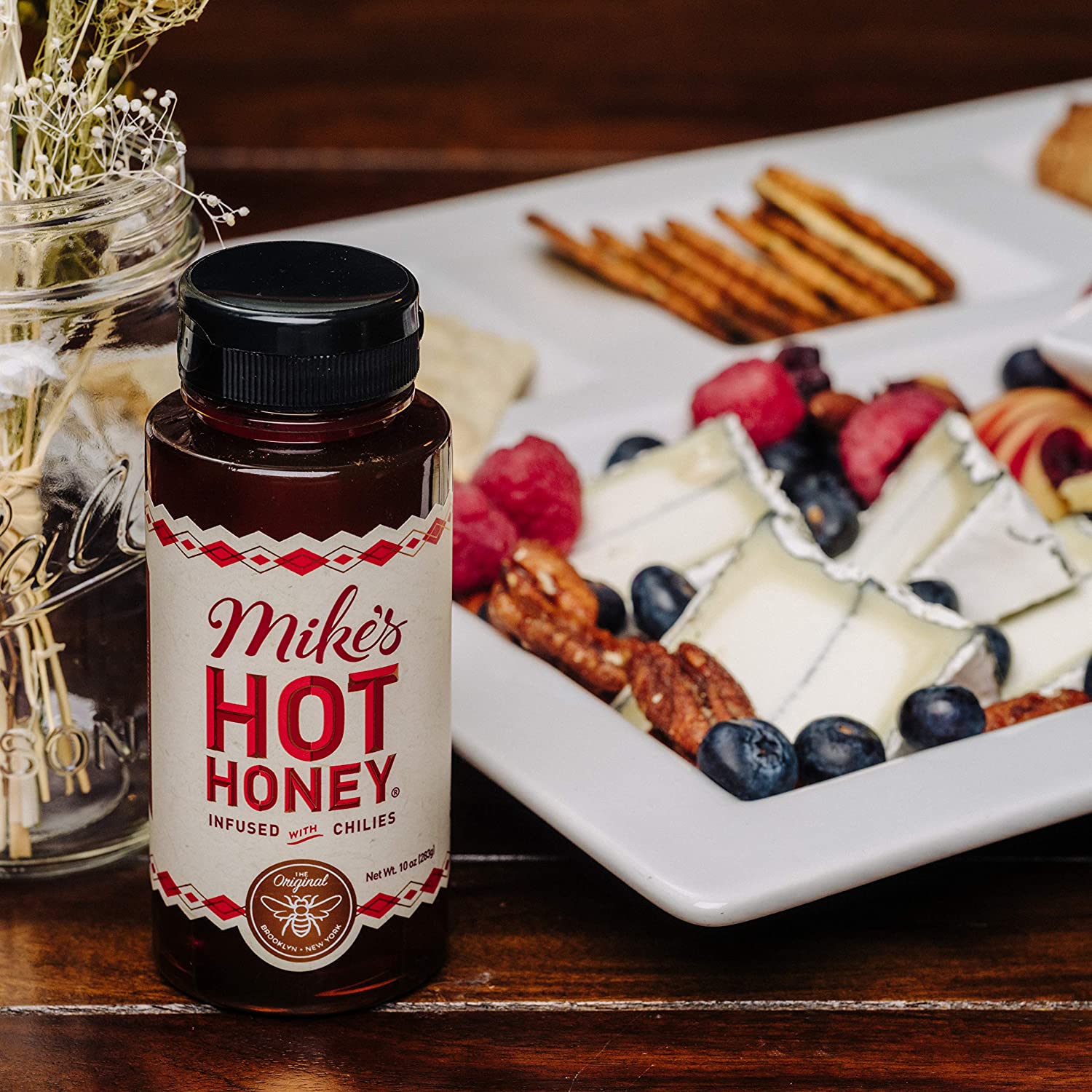 A bottle of Mike's hot honey is on a table next to a platter filled with cheese and cracker.