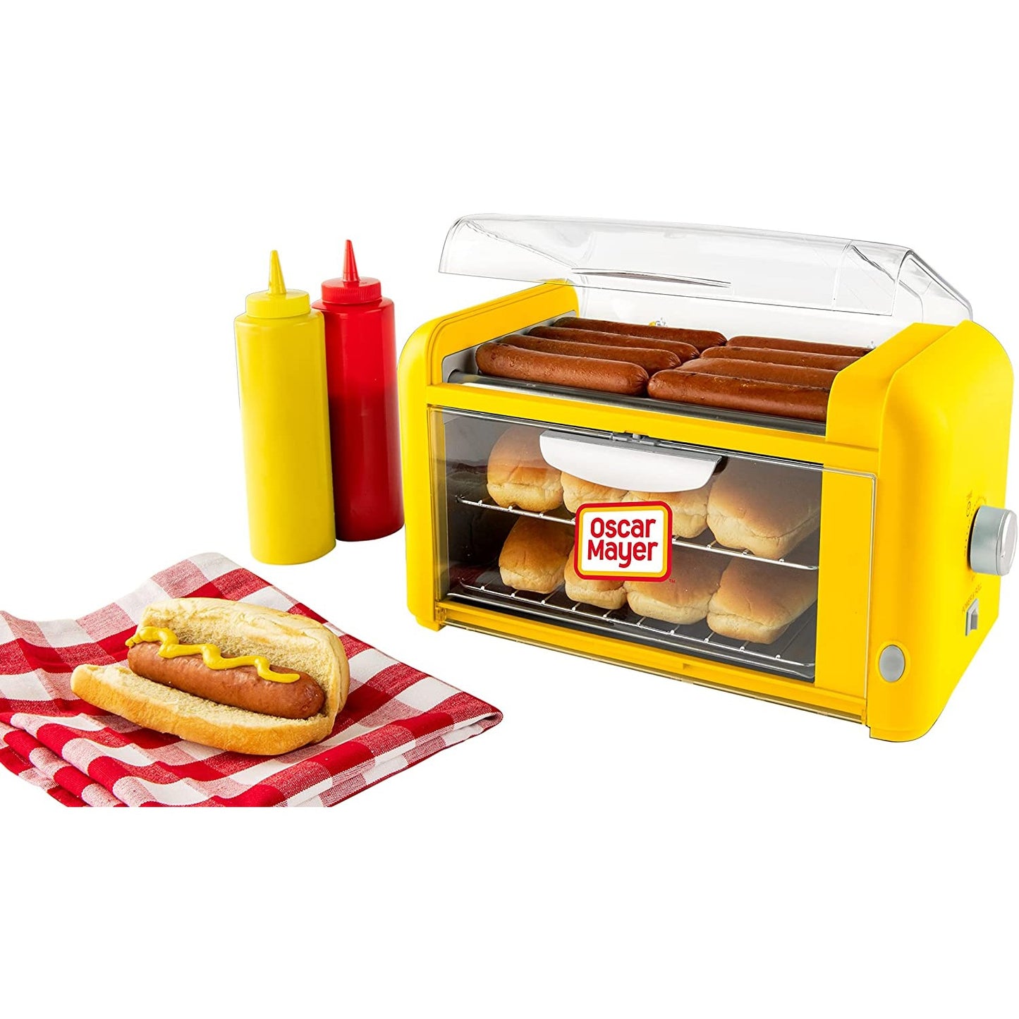 A hot dog toaster oven filled with sausages and buns. Nearby there are two bottles of sauce and a cooked hot dog on a napkin.