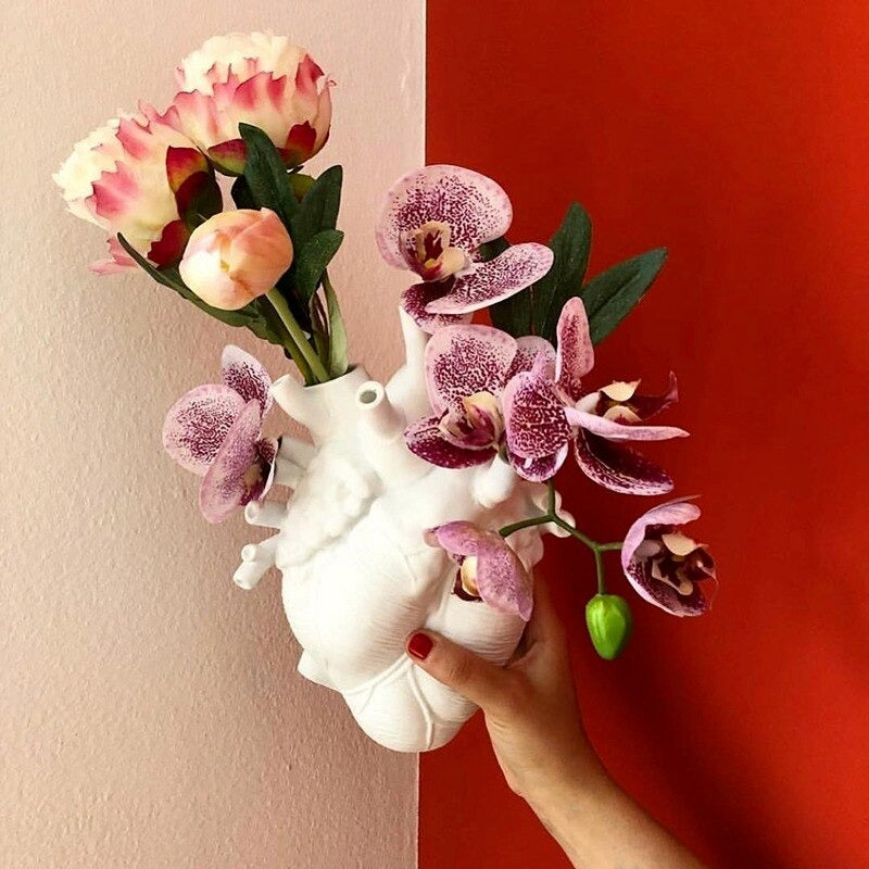 A hand is holding a white human heart shaped vase in front of a red and white background. The vase is filled with purple/white orchids.