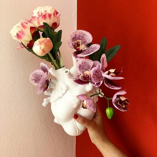 A hand is holding a white human heart shaped vase in front of a red and white background. The vase is filled with purple/white orchids.