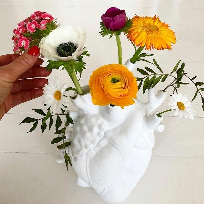 A white human heart shaped vase complete with arteries. The holes of the arteries are filled with various colored flowers and a human hand is placing a flower into one of the holes.