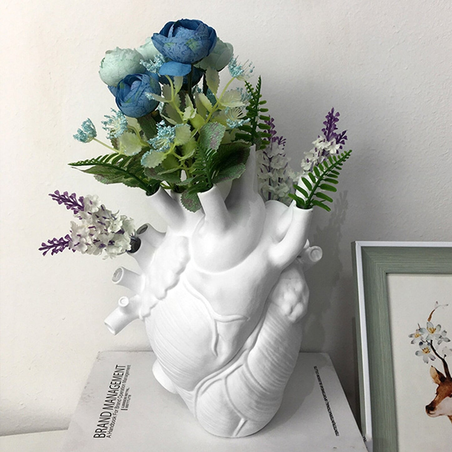 A white human heart shaped vase complete with arteries. The holes of the arteries are filled with various colored purple flowers and leaves. The vase is displayed resting on a book.