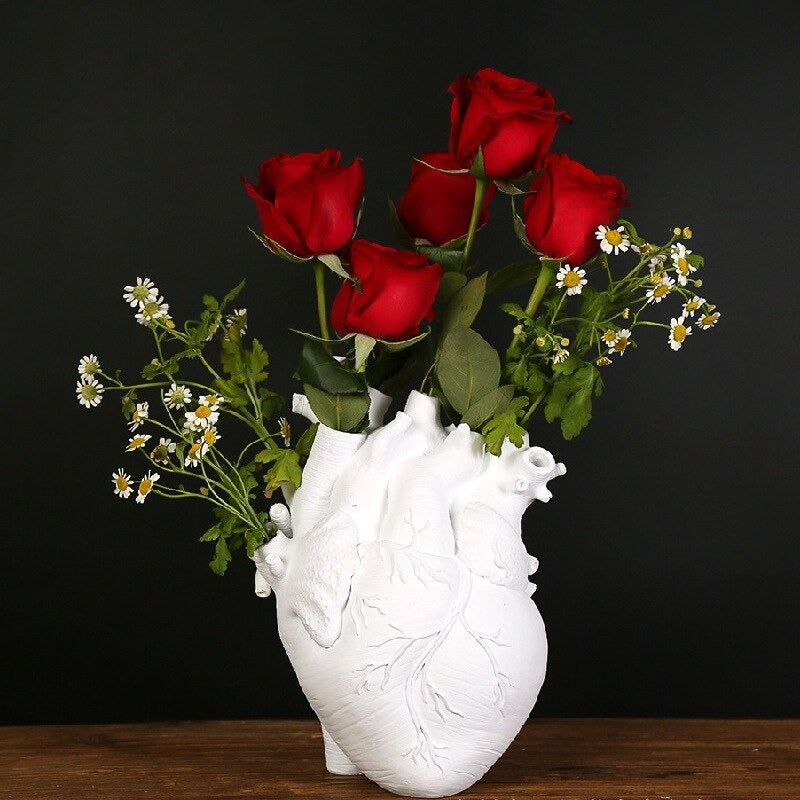 A white human heart shaped vase complete with arteries. The holes of the arteries are filled with red roses and tiny white daisies. The vase is on a wooden table.