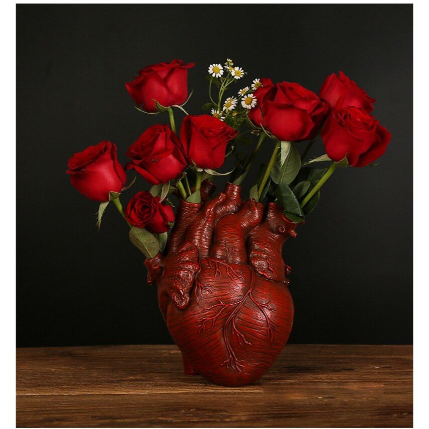 A blood red colored human heart shaped vase complete with arteries. The holes of the arteries are filled with red roses and tiny white daisies. The vase is on a wooden table.