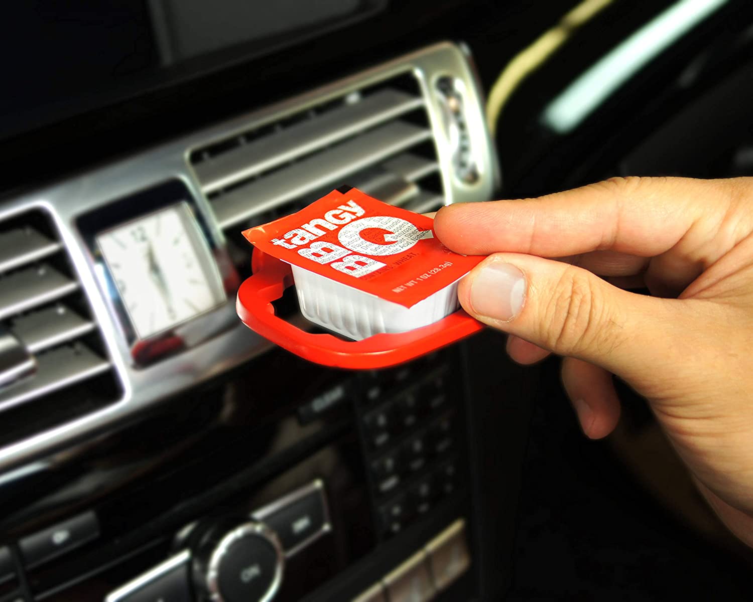 A sauce holder which is plugged into the vent of a car and is designed to hold your dipping sauce. A hand is placing some tangy BBQ sauce into the tray.