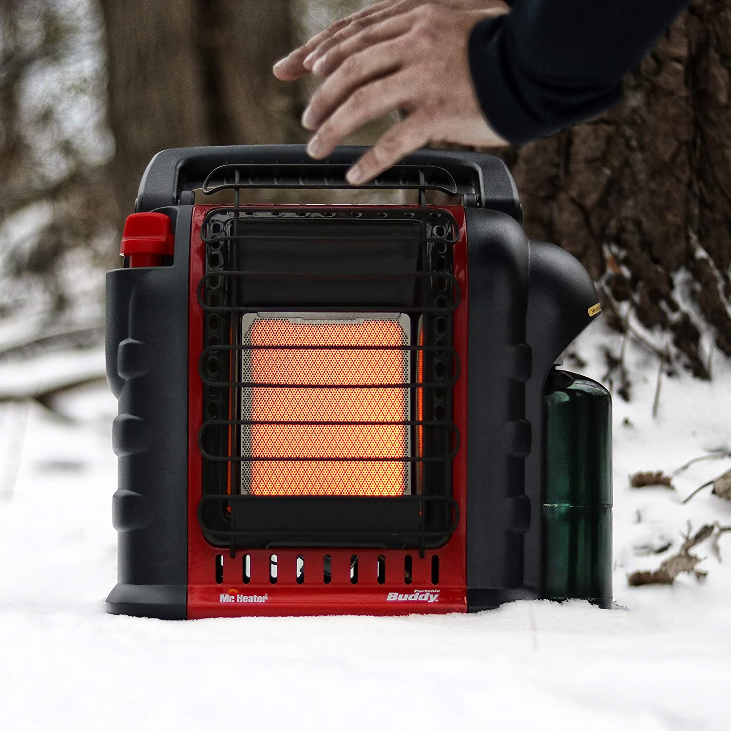 A person is warming their hands next to a propane radiant heater while outdoors in the snow.