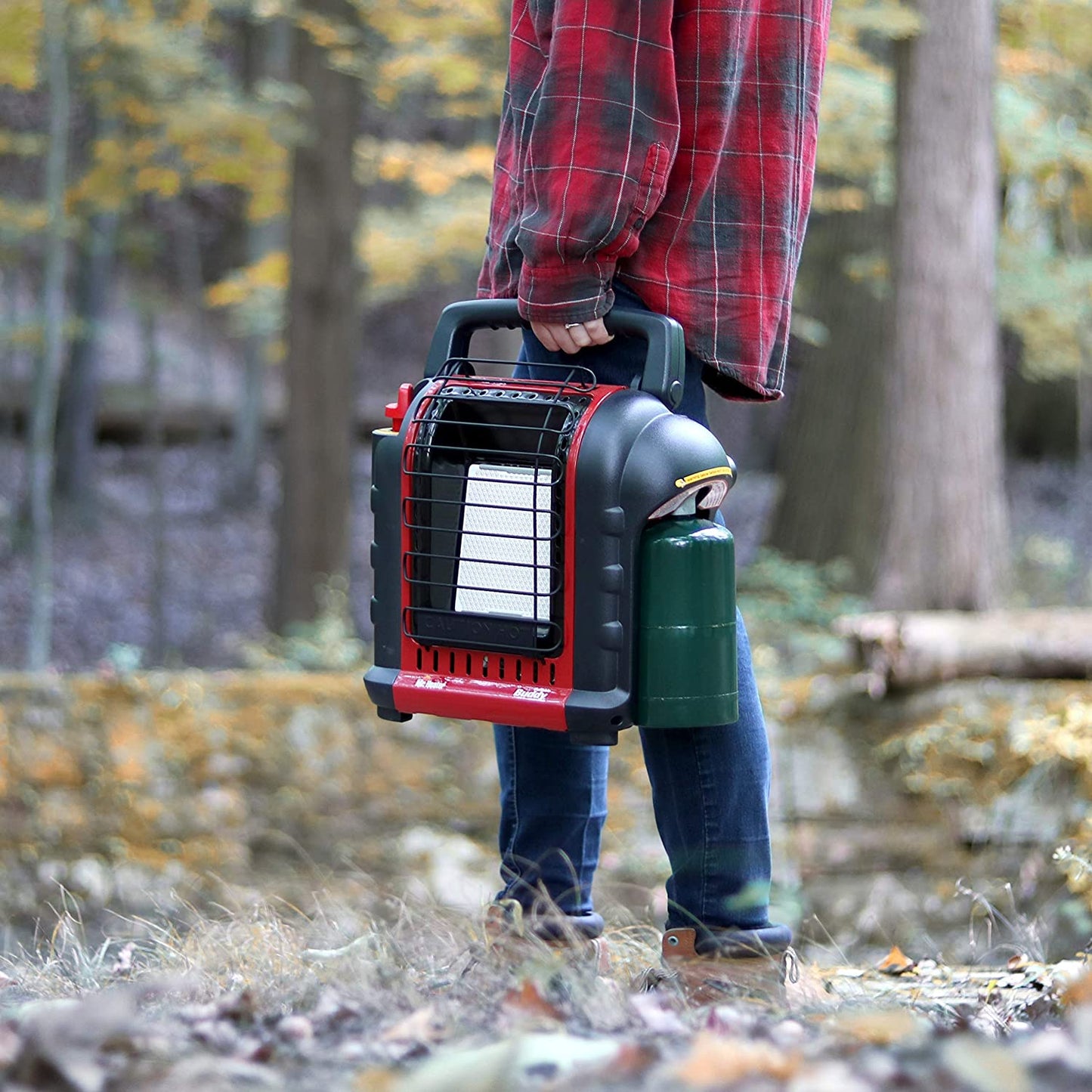A person is carrying a red and black propane radiant heater in the woods.