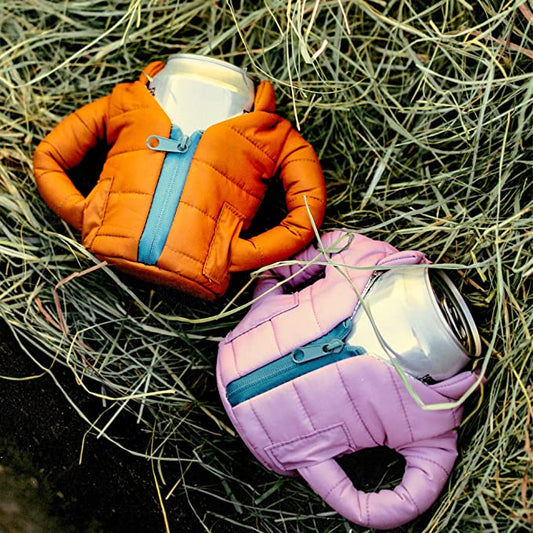 Two insulated drink cool jackets which looks like mini parkas. They each have a can of drink inside them and are laying on some dried grass.