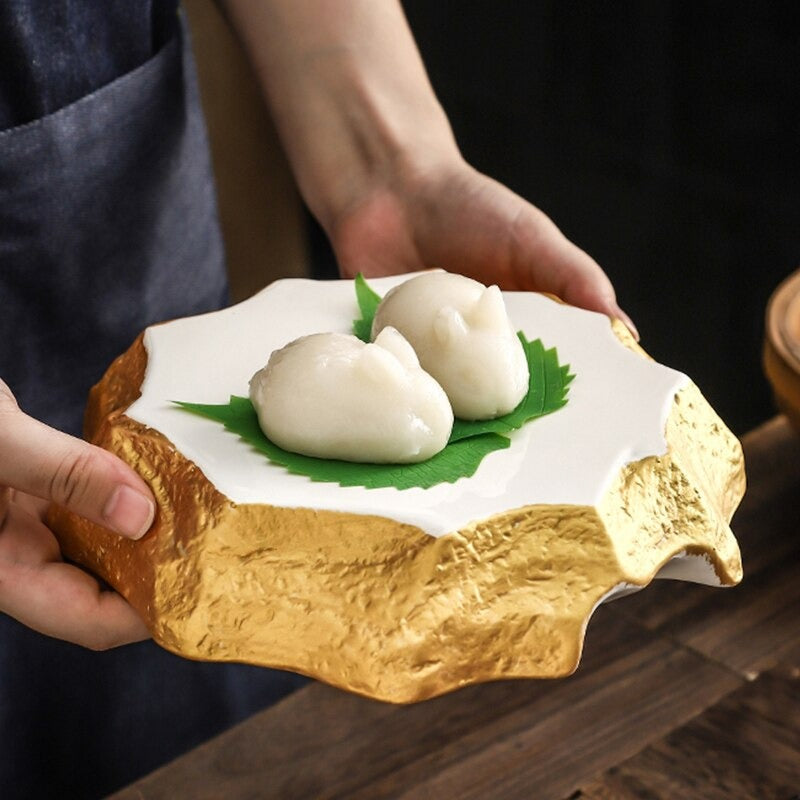 An irregular shaped ceramic dinner plate. The plate has gold colored sides and a white ceramic top. The plate is held by two hands and there are 2 Chinese dumplings on the plate.