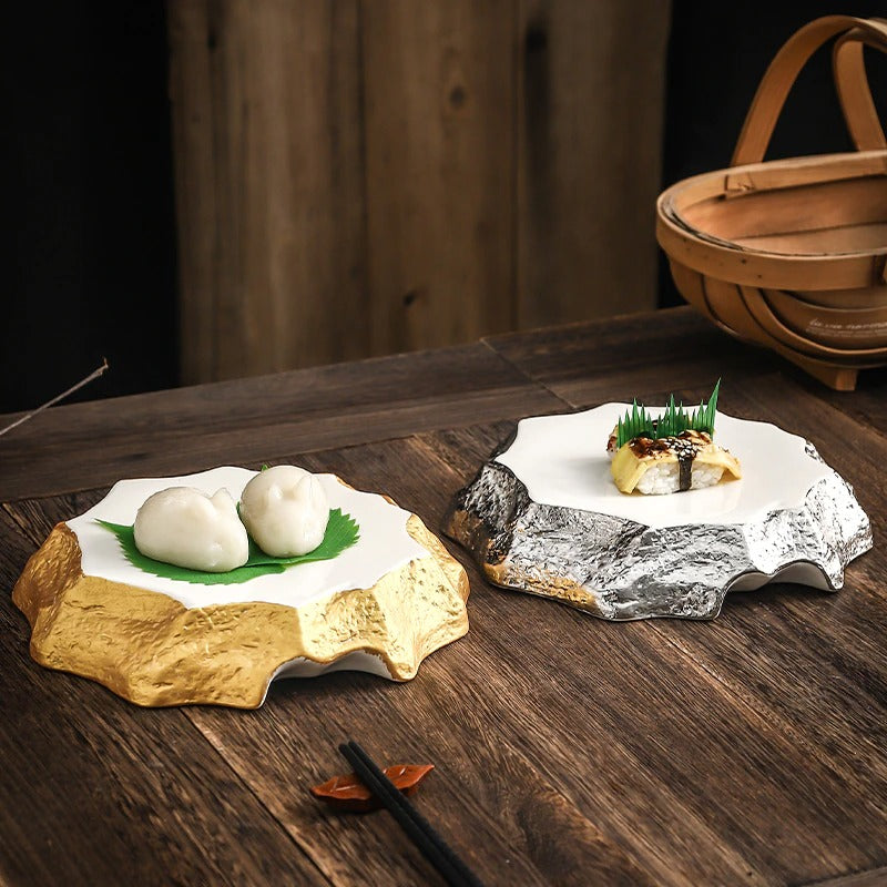 Two irregular shaped ceramic dinner plates. One is gold and one is silver. The gold plate has 2 Chinese dumplings on it while the silver plate has a piece of sushi on it.