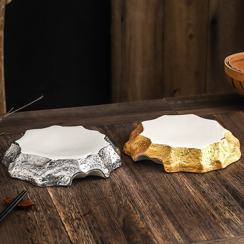 Two irregular shaped ceramic dinner plates. One has gold and one has silver edges and both have a white ceramic top. The plates are on a wooden table.