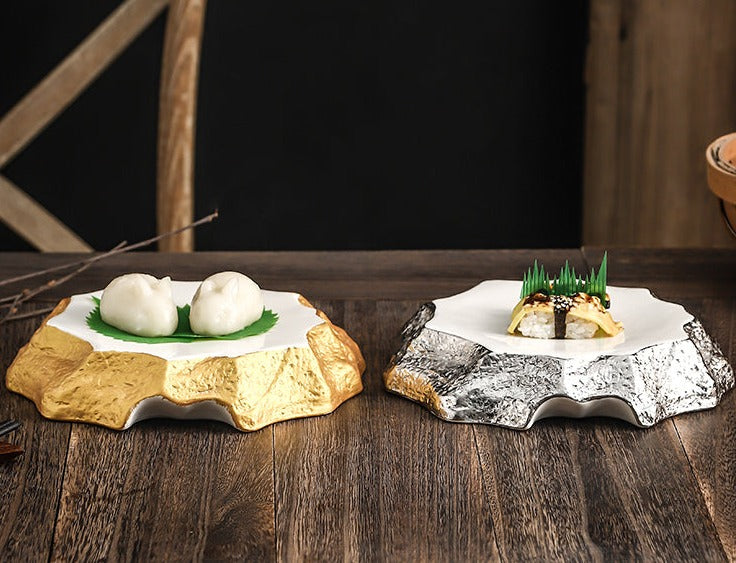 Two irregular shaped dinner plates. One plate has gold sides with a white ceramic top and has two Chinese dumplings on them. The other plate has silver sides with a white ceramic top and has sushi on it.