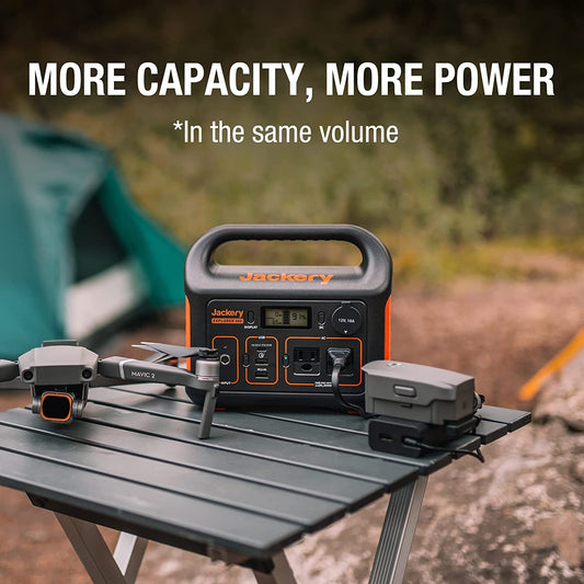 A Jackery portable power station is outdoors on a small table. There is a drone next to the power station. The text reads, 'More capacity, more power.'