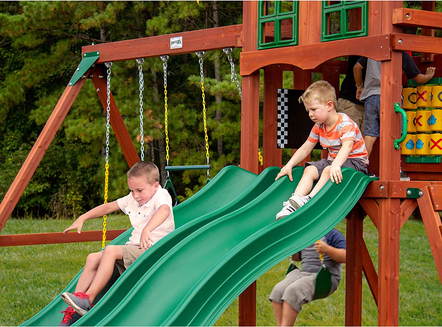 Two children are sliding down a pair of wave slides which is part of a large outdoor wooden play-set designed for kids.