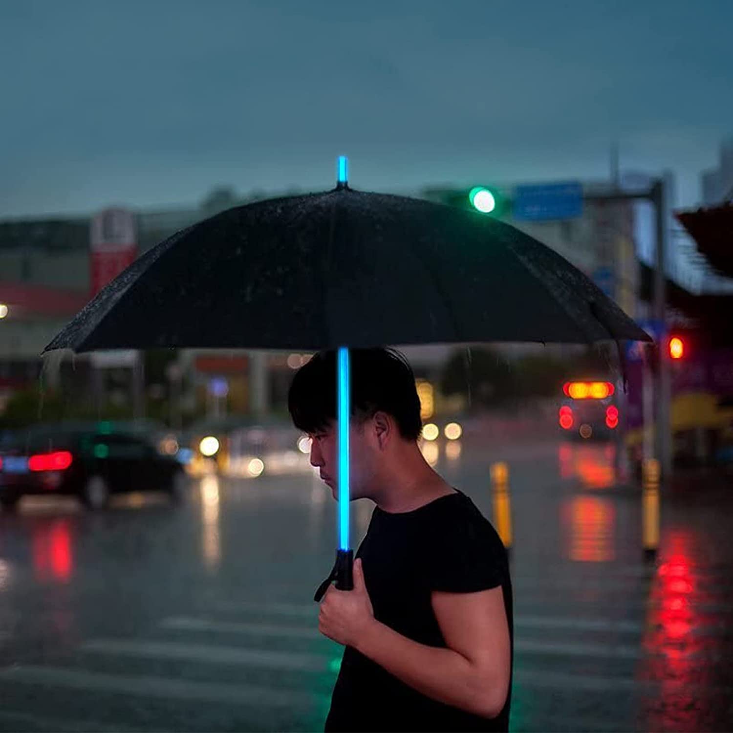 A person is walking outdoors holding a black umbrella which features a blue handle shaped like a lightsaber