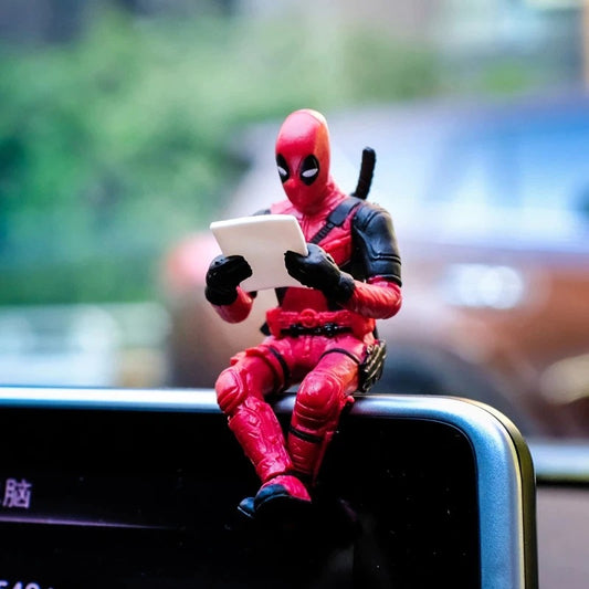 A Deadpool action figure sitting on a car dashboard. Deadpool has his legs crossed and looks as if he is reading something.