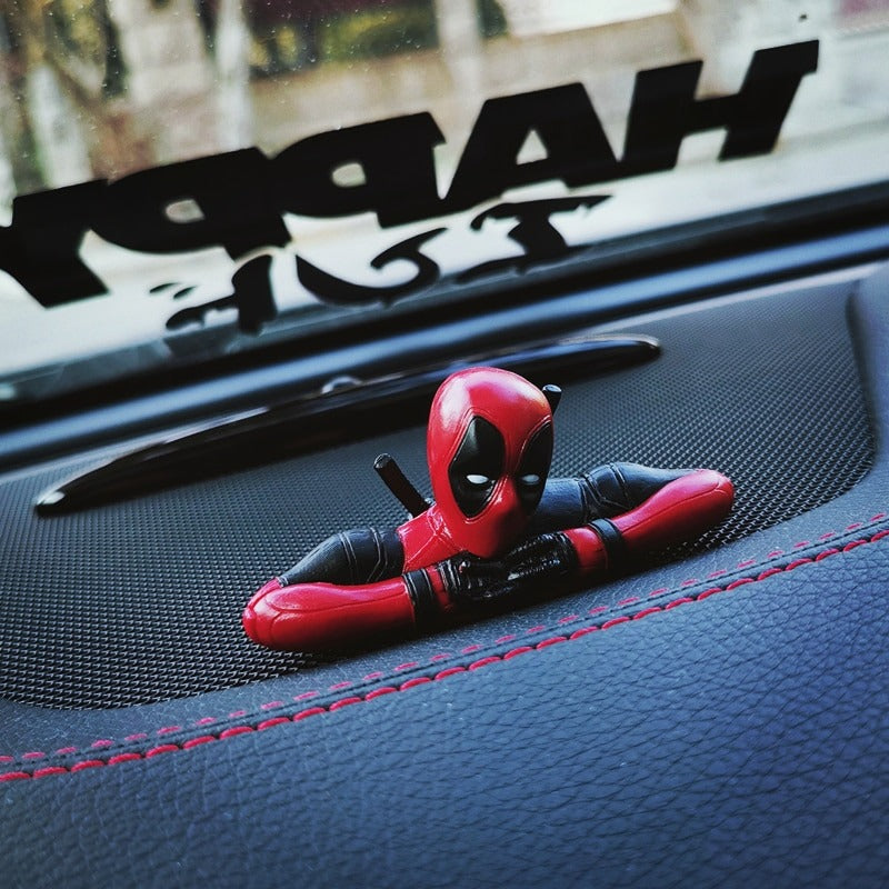 A head and shoulders only Marvel Deadpool action figure. He has his fingers interlocked and is sitting on a car dashboard.