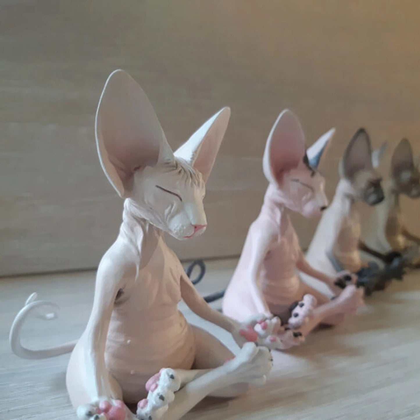 4 meditating Sphynx toy cats sitting in a yoga pose with their legs crossed and fingers pinched as if they are meditating. They are sitting in a row on the floor and are in various colors.