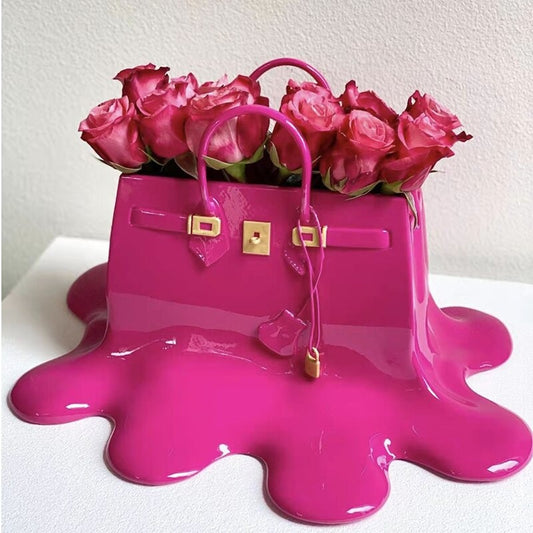 A pink resin vase which looks like a melting designer women’s handbag. The vase is filled with a bouquet of pink roses.
