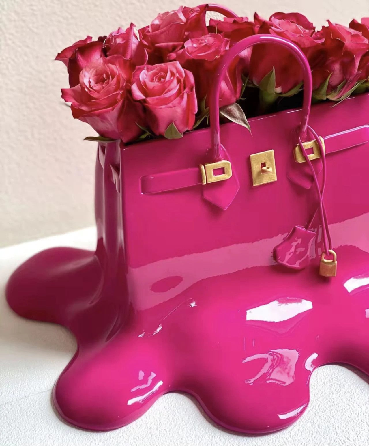 A close up view of a pink colored resin vase which looks like a melting designer women’s handbag. The vase is filled with pink roses.