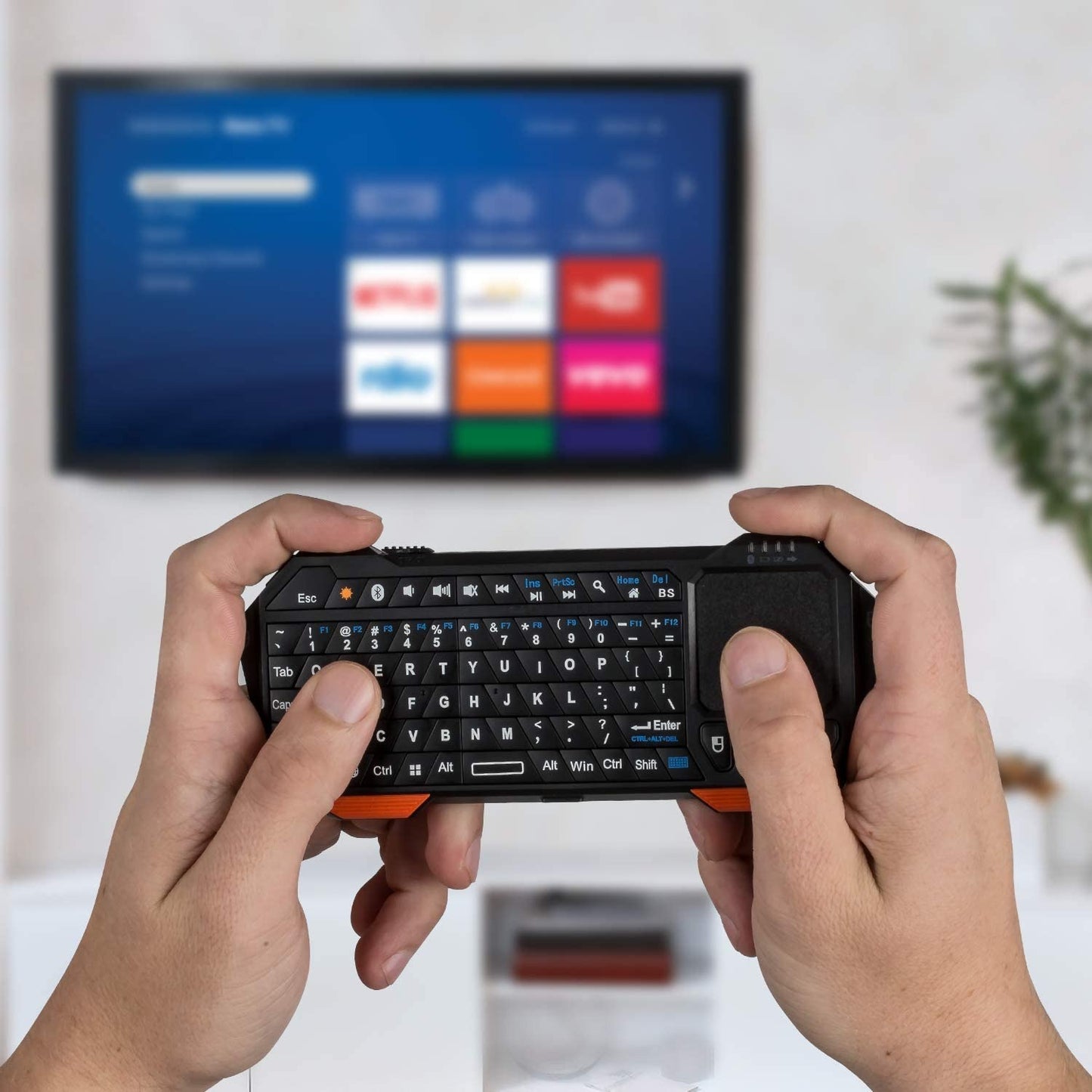 A pair of hands are holding a black colored mini Bluetooth keyboard with touchpad in front of a TV.