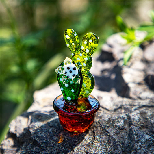 A small green and brown colored glass cactus figurine resting on a rock outdoors.