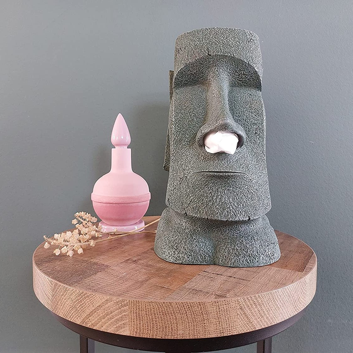 A moai shaped tissue box holder on a table next to a pink bottle.