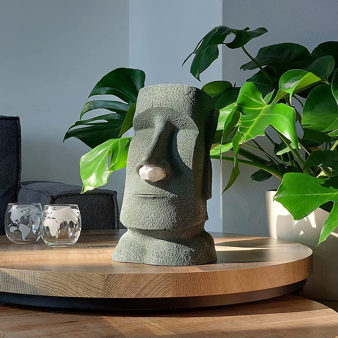 A moai shaped tissue box holder on a wooden table with a green plant behind it.