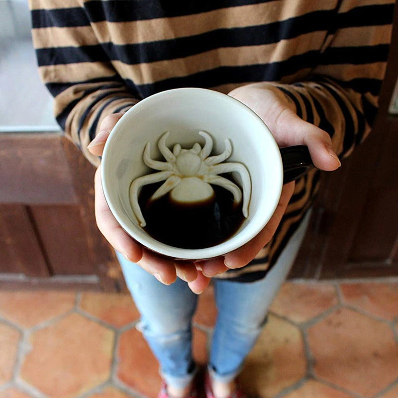 A person is holding a white mug which has a hidden white fake spider at the bottom of it.