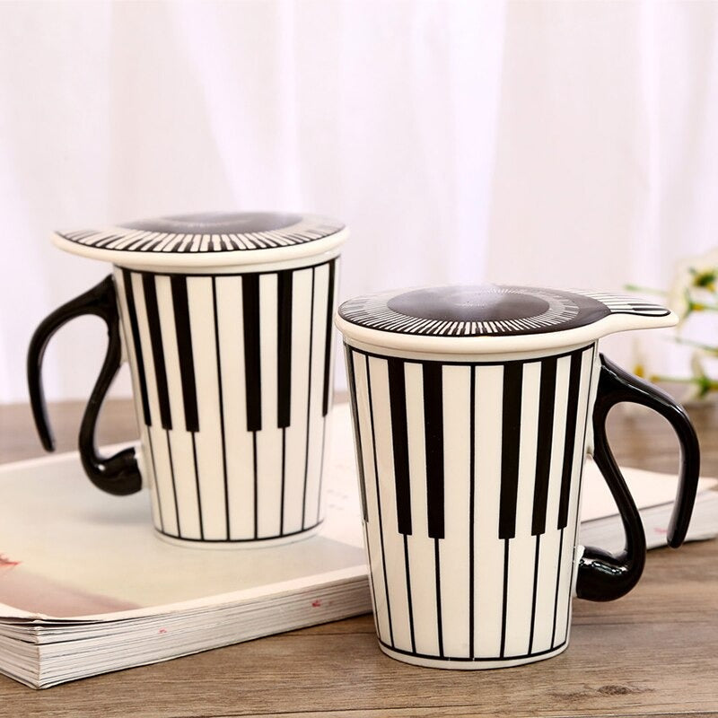 Two music mugs with lids. They two mugs and lids have a piano keyboard printed on them and both feature black handles.