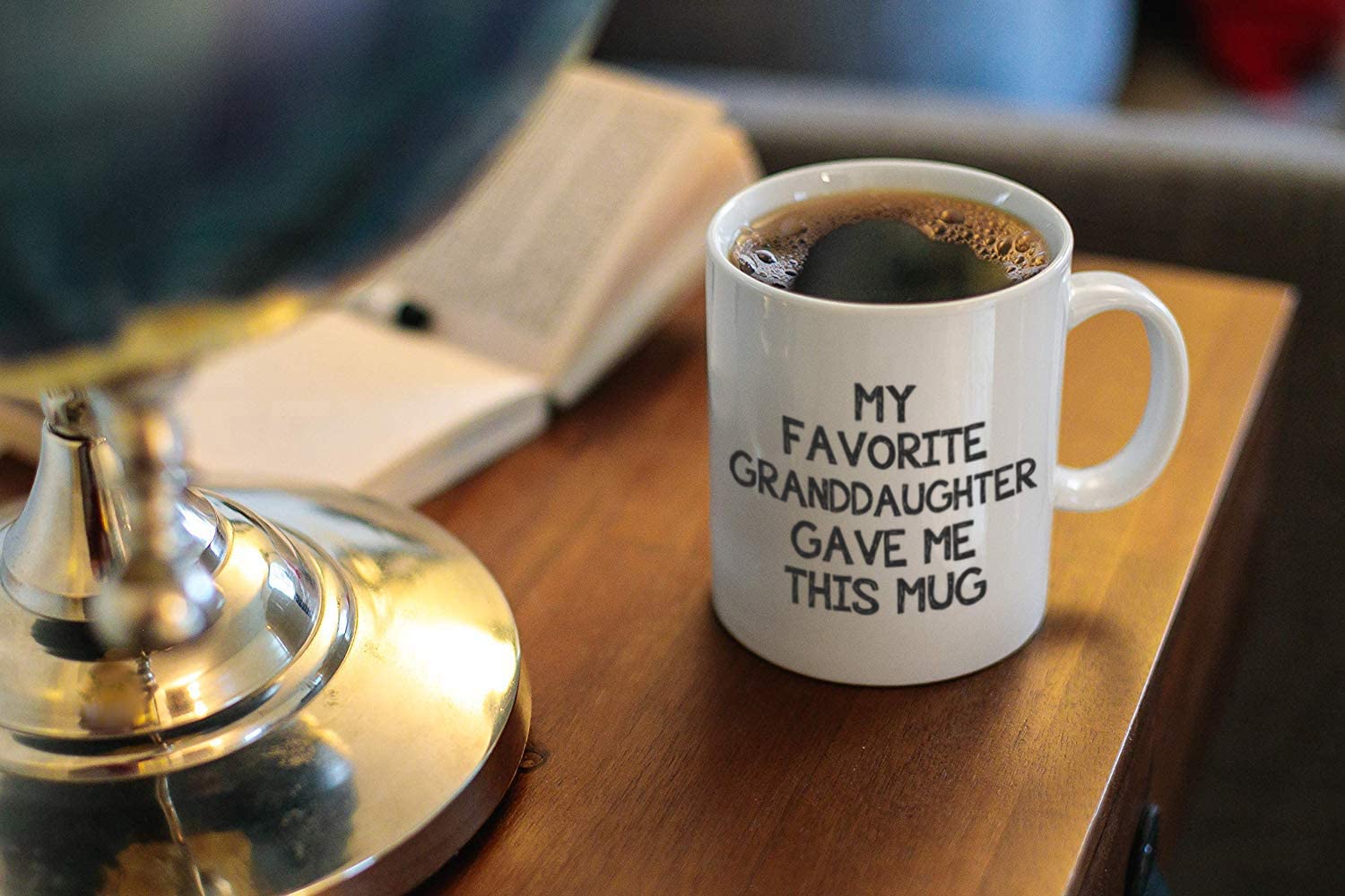A white mug full of black coffee on a wooden table. On the mug there are words printed on it which say, 'my favorite granddaughter gave me this mug'