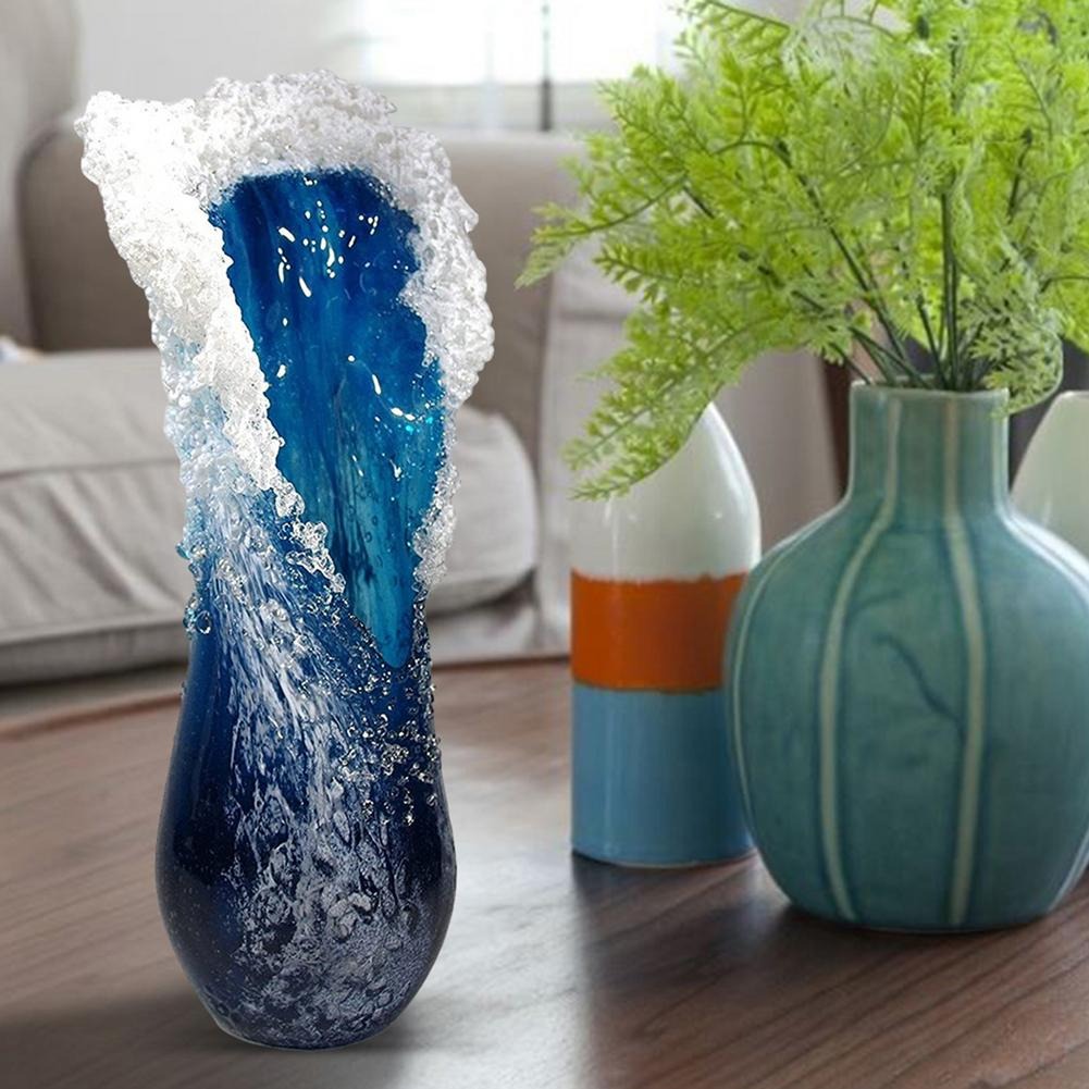 A blue and white ocean wave vase which looks like an ocean wave frozen in time. The vase is on a table next to 2 other vases one of which has some plastic ferns in them.