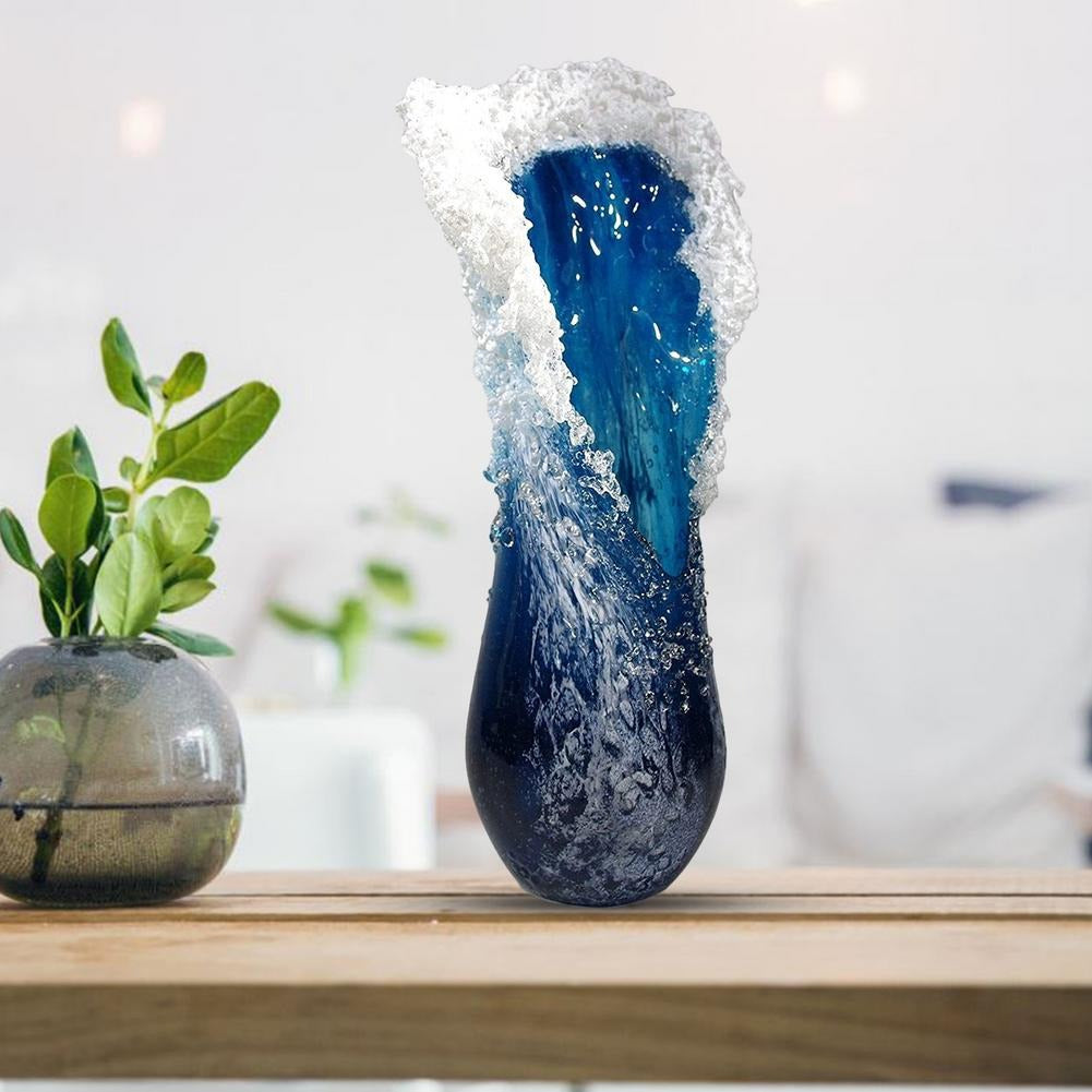 A vase which looks like a frozen ocean wave caught in time. The vase is blue and white and sits on a wooden table. Next to the ocean wave vase is a smaller ball shaped vase with some small leafy branches in it.