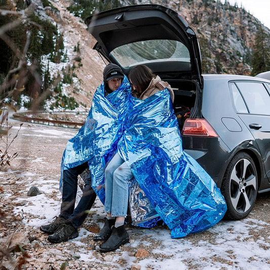 Two people are outside in cold, snowy weather. Each person has a blue emergency blanket wrapped around them.
