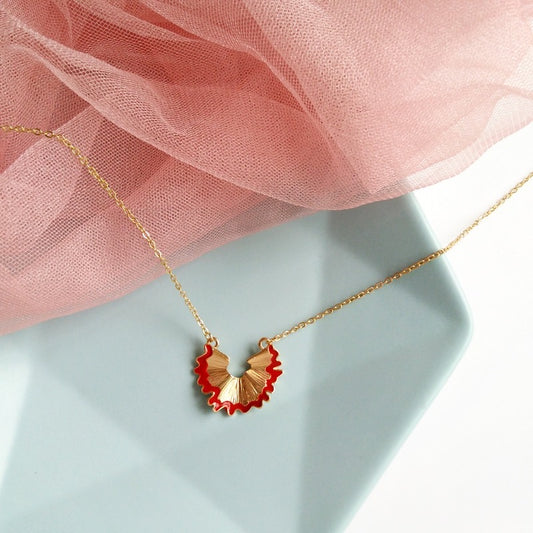 A pencil shaving necklace which features a gold chain and the gold pendant is in the shape of a red pencil shaving.