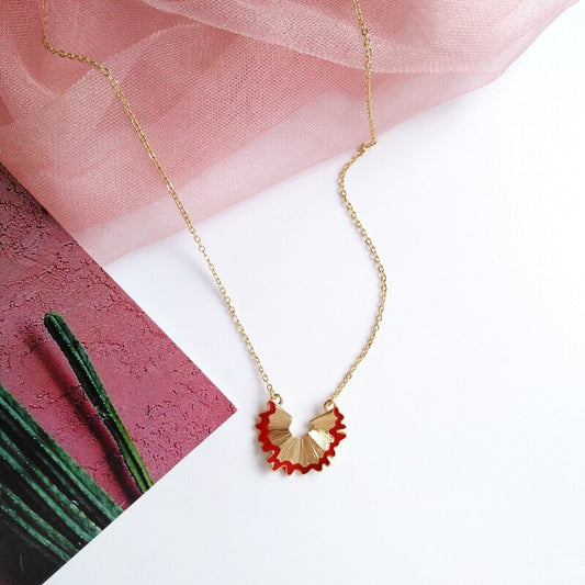 A gold necklace which features a pendant in the shape of a red pencil shaving.