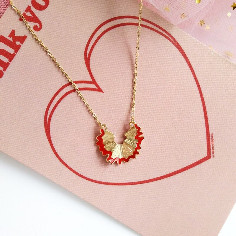 A red love heart is drawn around a pencil shaving necklace. The gold necklace features a pendant which has a pencil shaving shaped pendant on it.