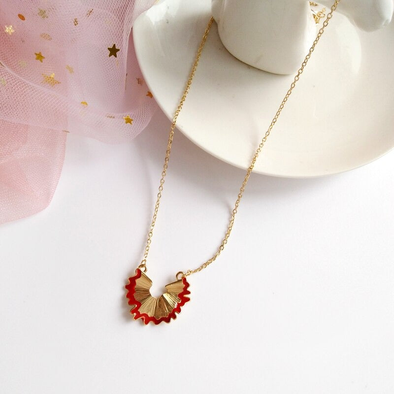A gold and red pencil shaving necklace which features a gold chain and a gold pendant in the shape of a red pencil shaving.