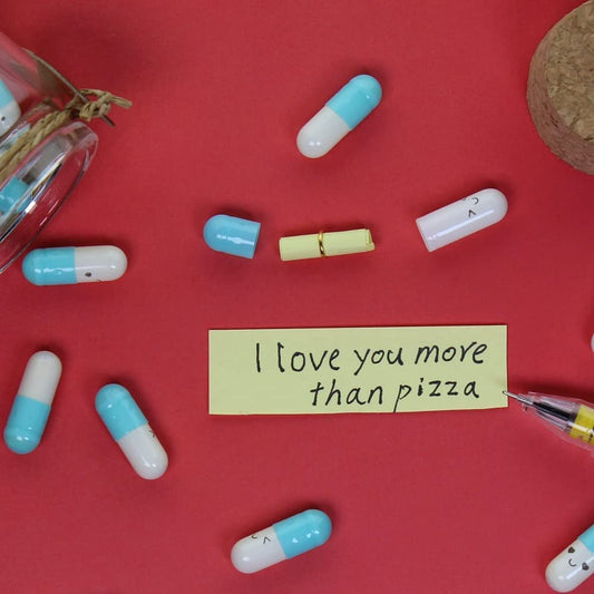 Light blue and white pill shaped capsules on a red background. One of the capsules is open and the love note has been revealed, it reads, "I love you more than pizza."