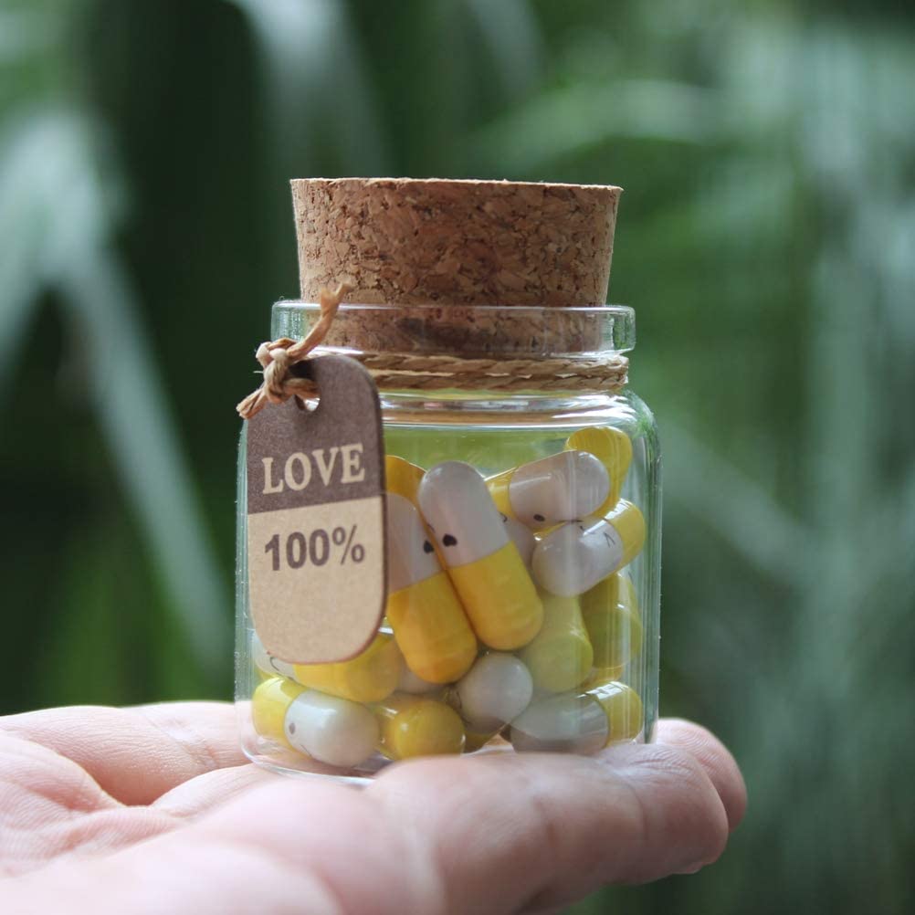 The palm of a hand is holding a small glass bottle with little yellow and white pill shaped capsules inside. Each capsule contains a love note. There is a tag on the glass bottle which reads, '100% love.'