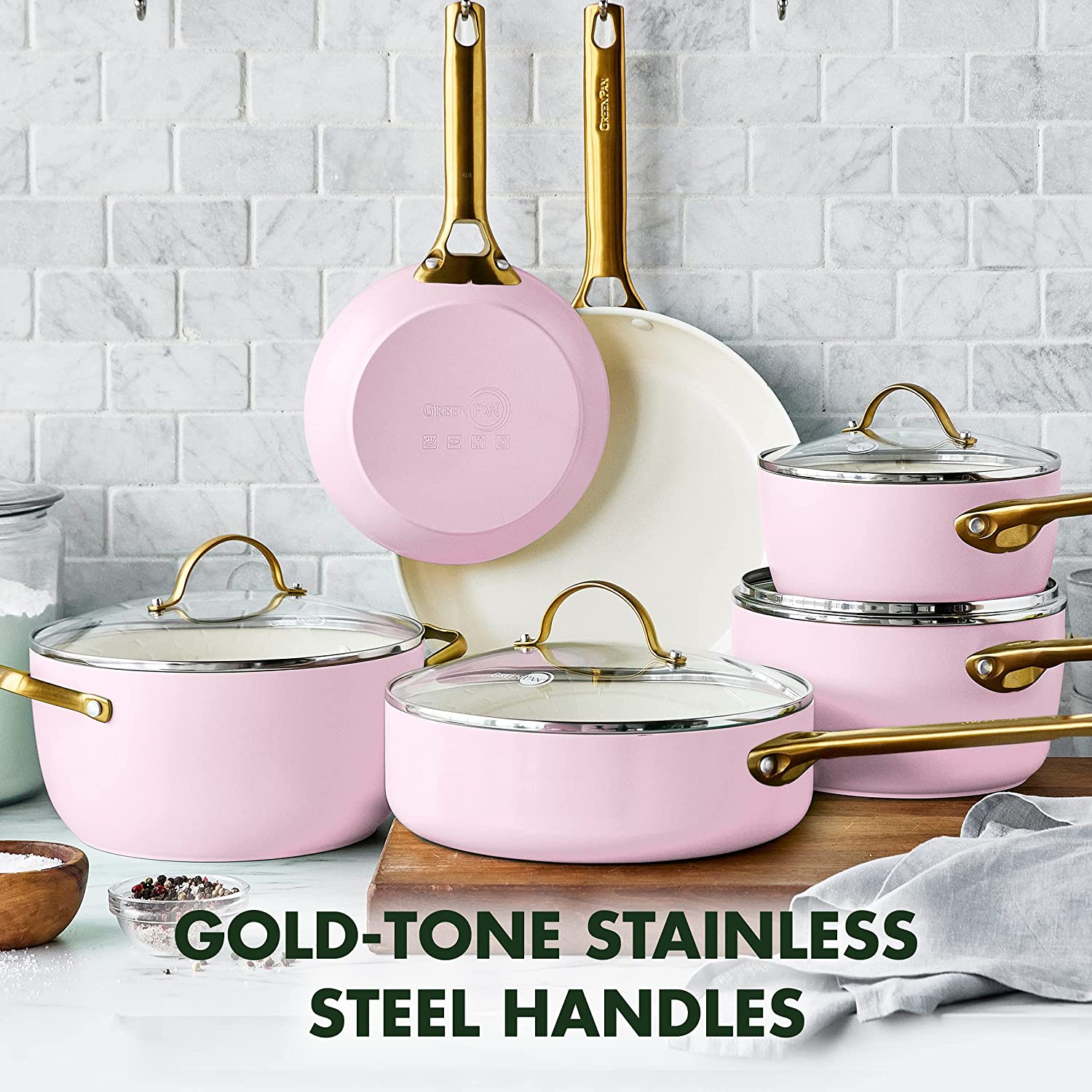 A pink cookware set comprising of pots and pans are in a kitchen. The headline says, 'Gold-tone stainless steel handles.'