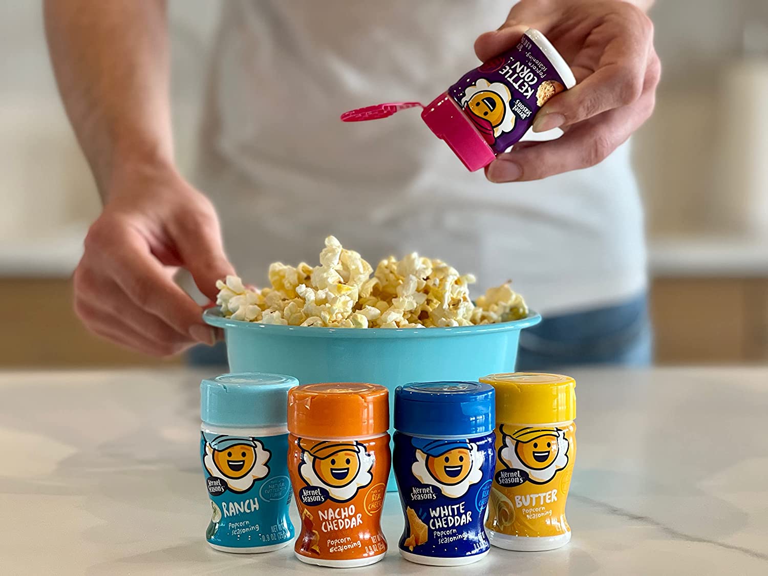 4 different flavored popcorn seasoning jars. Someone is using a fifth jar to sprinkle some seasoning on freshly cooked popcorn.