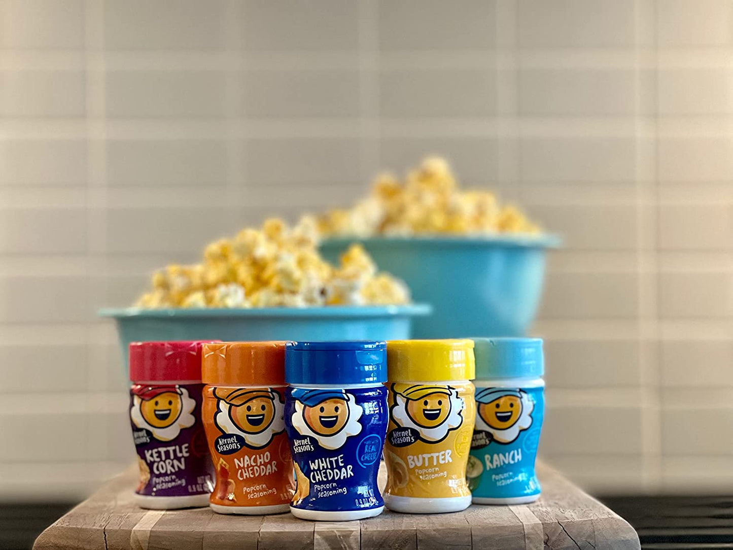 Five various flavored popcorn seasoning jars with two bowls of fresh popcorn in the background.