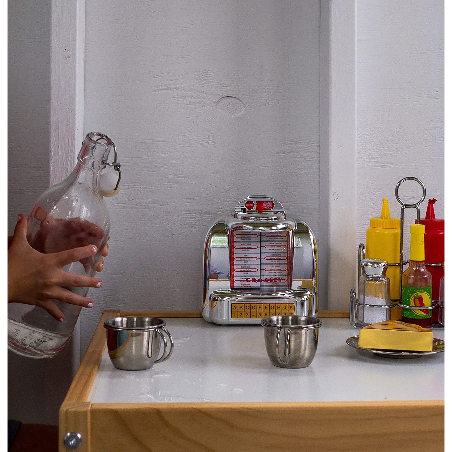 A retro radio which looks like a diner jukebox is on a kitchen bench with kitchen accessories surrounding it.