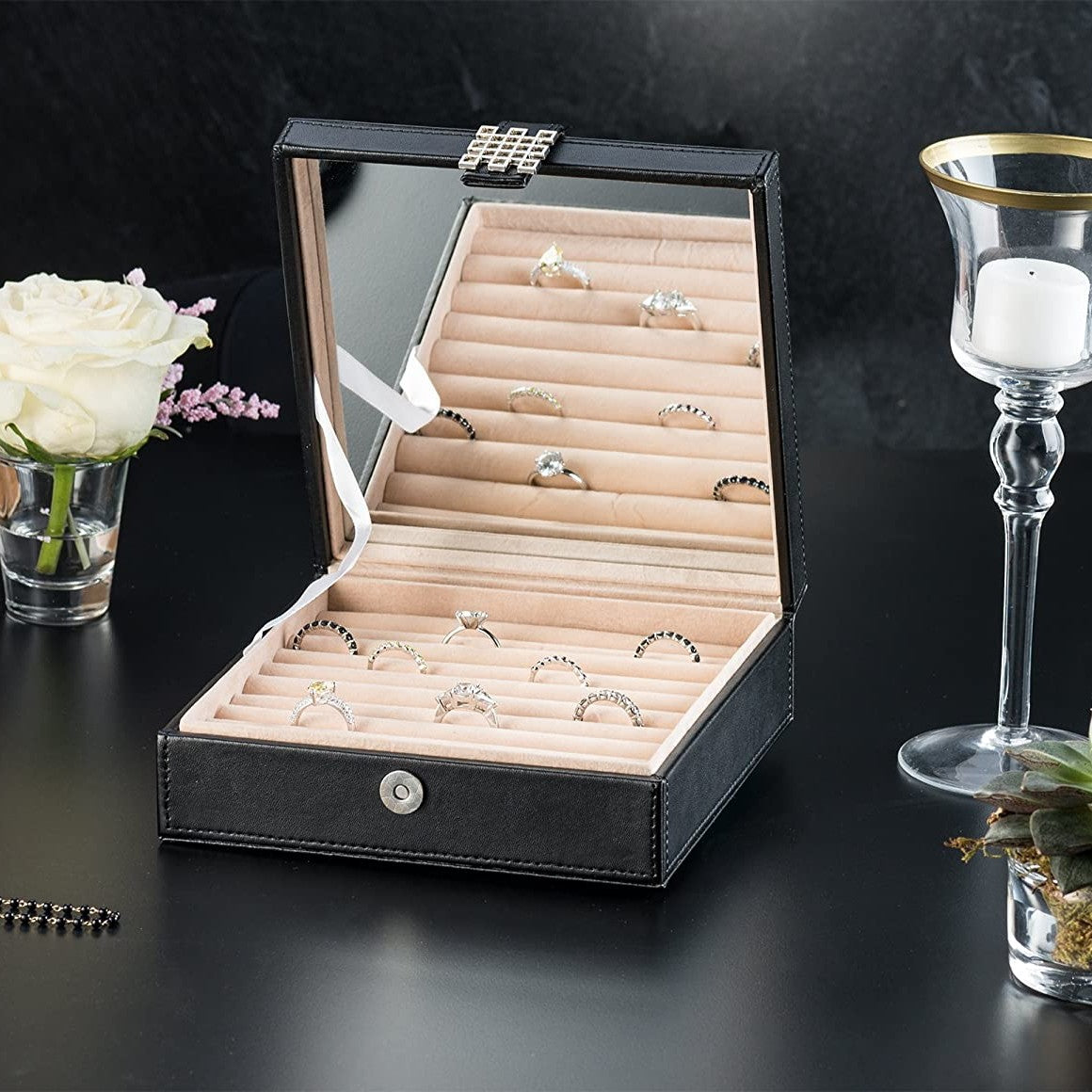 A stylish looking black ring box organizer with several rings inside and the lid is open. There is a candle in a glass and a white rose nearby.