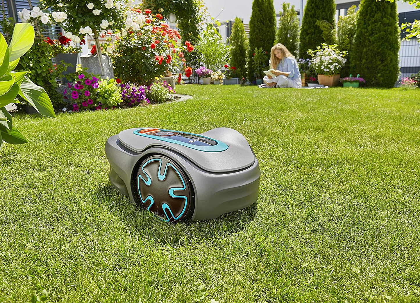 A robotic lawn mover on a green lawn with a woman reading a book in the background.