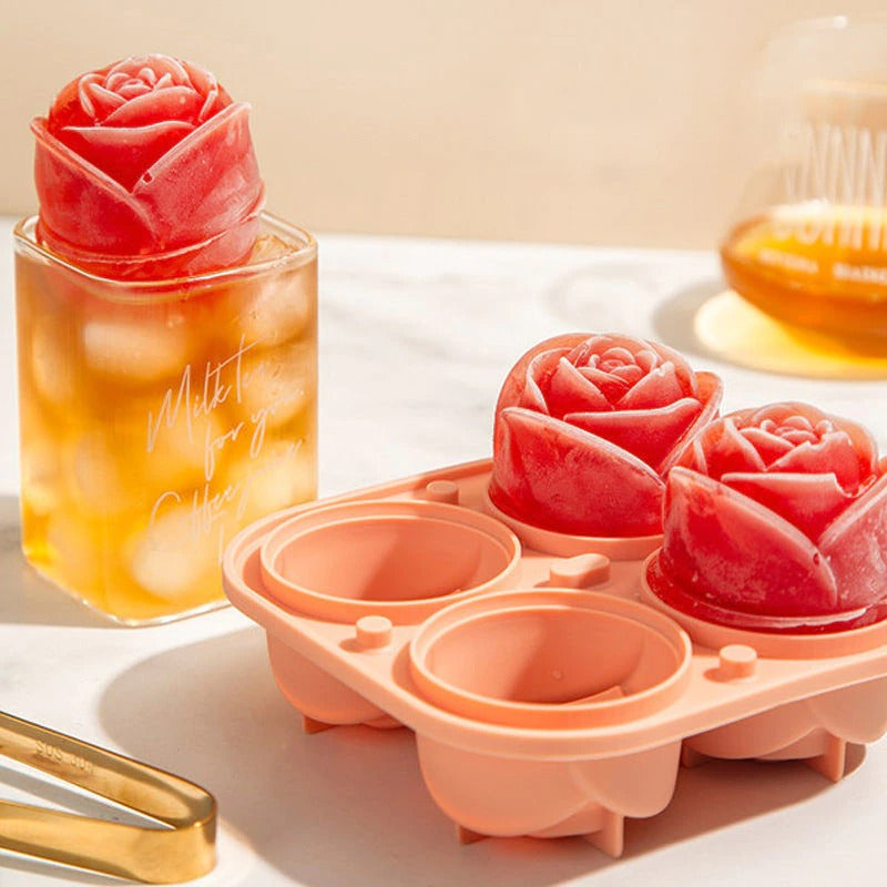 An ice cube mold which creates rose shaped ice cubes, there are two red colored rose shaped cubes in the tray and a drink nearby which has a red colored rose shaped ice cube in it.